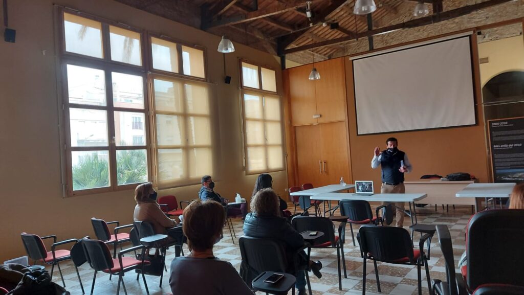 Sala amb sostre de bigues de fusta i una pantalla de projecció, on es veu un ponent parlant a un grup reduït de persones assegudes.