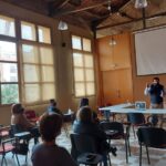 Sala amb sostre de bigues de fusta i una pantalla de projecció, on es veu un ponent parlant a un grup reduït de persones assegudes.